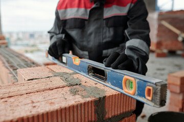 Gloved hands of male constructor or foreman in workwear holding level on surface of brick wall at...