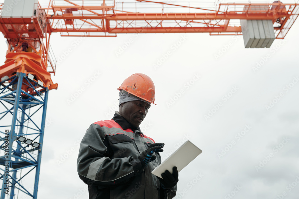 Poster mature black man in gloves, workwear and hardhat scrolling through online information while using ta