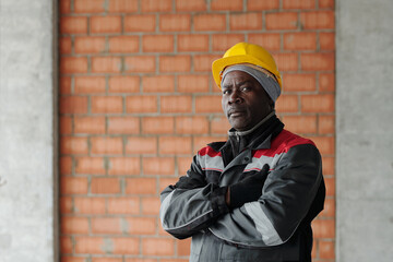 Modern African American male worker of construction site looking at you while standing against brick wall and keeping arms crossed on chest