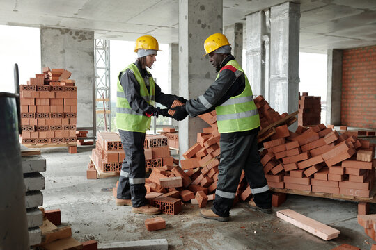 Two intercultural colleagues in workwear preparing building materials for construction work while black man passing bricks to young female