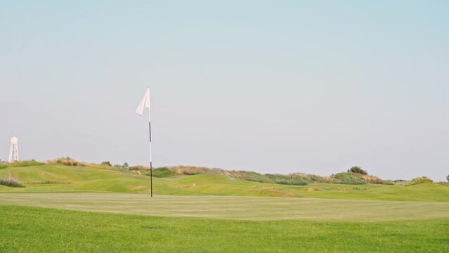 Static meditative footage. A white flag on a golf hole flutters in the wind. Green bright grass on the golf course against the blue sky