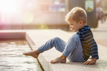A cute little boy, barefoot, wants to climb into the water at the city fountain during the summer.