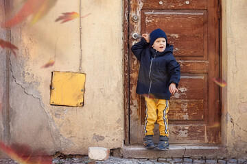 A little boy poses on an autumn day at the ancient door and cracked wall in the Old Town. The autumn wind blowing up the colorful leaves of the trees, child is surprised by the fall of leaves