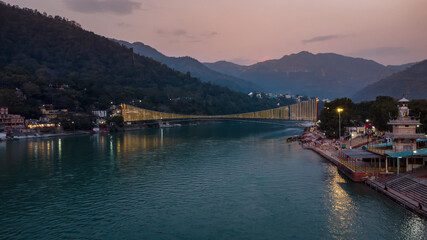 Fototapeta na wymiar iron suspension bridge illuminated with ferry lights with ganges riverbank view at evening aerial