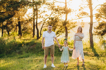 Fototapeta premium Young beautiful family with a little daughter hug, kiss and walk in nature at sunset. Photo of a family with a small child in nature.