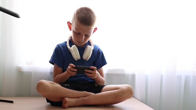 Child's dependence on a mobile device theme. Child with headphones on neck is sitting on table and uncontrollably playing online games on smartphone. virtual life. children's addiction to online games