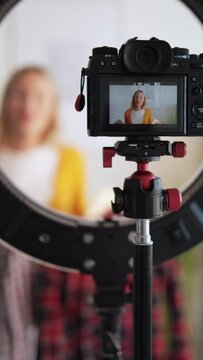 Fashion blog. Female stylist. Video record. Smiling woman showing two checkered shirts on camera screen in light home studio interior vertical.