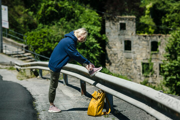 A female hiker with a yellow backpack is tying her shoelaces on the road.