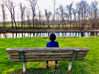  Boy sitting on a park bench looking at a Serene river. calm, peaceful, and undoubtedly tranquil