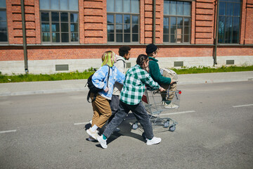 Group of teenagers playing outdoors together, they riding each other on shopping cart along the street