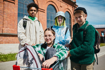 Fototapeta na wymiar Portrait of group of teen boys and girls looking at camera while riding on shopping cart on the street