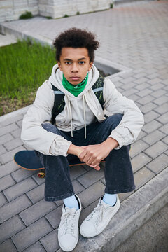 Portrait of African teenage boy sitting on his skateboard and looking at camera spending time outdoors