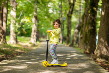 A small, beautiful girl, in a yellow sweater and sweatpants, rides a scooter in the park.