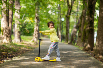 A small, beautiful girl, in a yellow sweater and sweatpants, rides a scooter in the park.