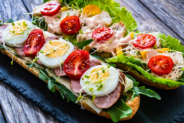 Long sandwiches with sausage, ham, chicken breast, boiled eggs, lettuce, arugula yellow cheese, tomato and dressing on wooden table
