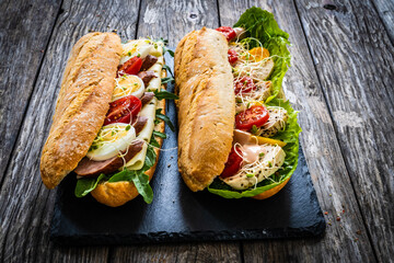Long sandwiches with sausage, ham, chicken breast, boiled eggs, lettuce, arugula yellow cheese, tomato and dressing on wooden table
