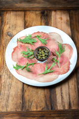 Marbled beef carpaccio with arugula and capers on white plate, wooden background, top view