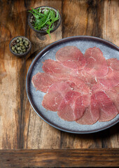 Marbled beef for carpaccio with ingredients arugula and capers on gray plate, wooden background, top view
