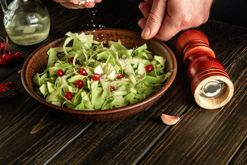 Professional chef sprinkles salt to salad of fresh vegetables on wooden table. Preparing healthy food on the kitchen