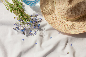White organic cotton blanket with forget me not flowers, glass of water and straw hat. Summer...