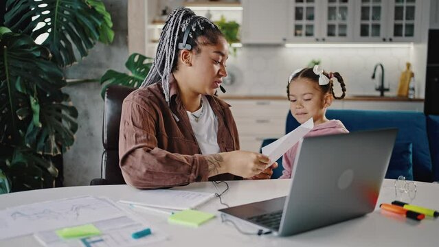 Work from home. Young black woman working remotely from home, little girl coming to mother and showing her picture