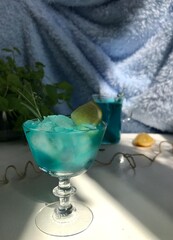 Blue cold drink in a glass with ice cubes on white desk with blue background. Also used mint leaves and rosemary, rope, spoon  with natual sunlight from the open window