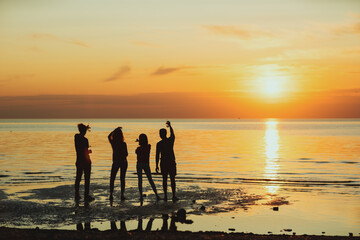 Silhouettes of four friends cheering with beer bottles standing in sea water on beach during...