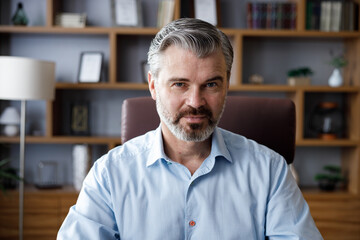 Obraz premium Business portrait of confident handsome bearded businessman looking at camera and smiling. Front close up portrait of Caucasian gray-haired man indoors at home office. Human face.