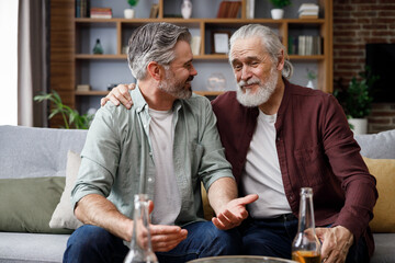Fathers day. Adult son with senior father drinking beer sitting on couch at home. Two generations...
