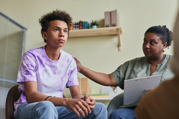African teenage boy talking during group therapy session with psychologist supporting him - obrazy, fototapety, plakaty