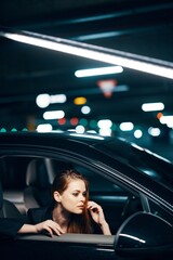 vertical photo from the side, at night, of a woman sitting in a black car and looking out of the window looking into the side view mirror holding her hand near her face