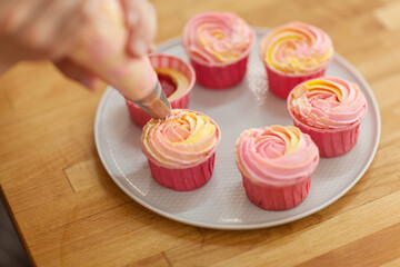 Obraz na płótnie Canvas Hands of pastry chef using piping bag to decorate delicious cupcakes with pink yellow buttercream frosting, from above closeup