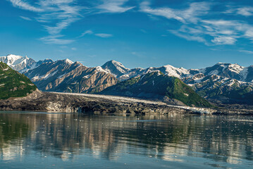 Disenchantment Bay, Alaska, USA - July 21, 2011: More dirt than ice at Turner Glacier landing under blue cloudscape. Snow topped mountain range on horizon and mirrored in ocean water.