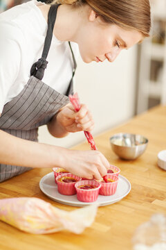 Side view of female confectioner using piping bag to fill freshly baked cupcakes with berry ganache frosting standing at cooking table in kitchen