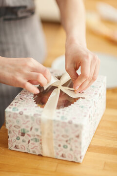 Hands of unrecognizable female pastry chef packaging cupcakes into gift box and tying it with ribbon, closeup