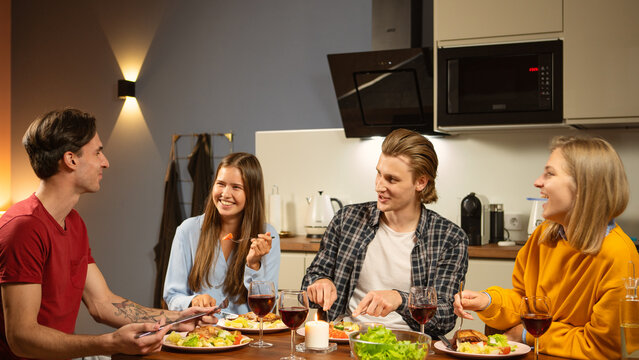 Group of four smiling and laughing young friends enjoying food and joyful talk sitting at table during home dinner with wine