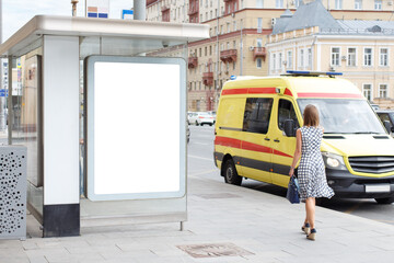 Advertising billboard vertical at the bus stop. An ambulance is on the road, a girl with a bag is walking along the sidewalk. Mock-up.