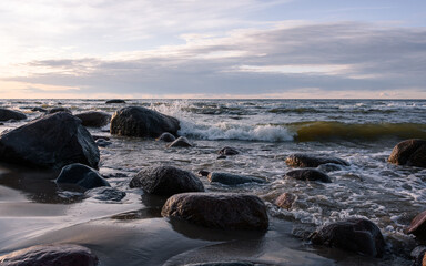 Fototapeta na wymiar Sea foaming waves crash on granite blocks, raising splash. Partly cloudy evening before sunset.