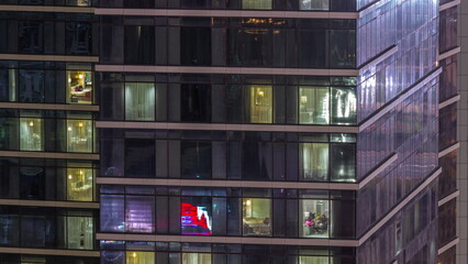 Fototapeta na wymiar Apartment windows of a glazed skyscraper glow at night with city lights reflection aerial timelapse.
