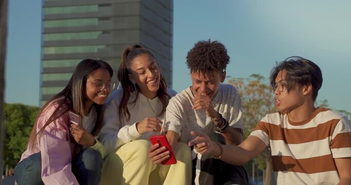 Group of multicultural happy teenage friends looking the phone and laughing in a bench in the city street.