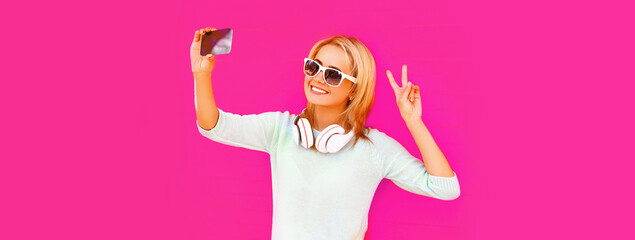 Portrait of happy smiling young woman taking selfie with smartphone in headphones listening to...