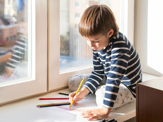 Little boy sits on window sill and draws rainbow with colored pencils. Creative leisure activity for children at home.