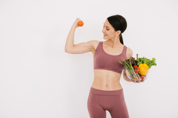 Beautiful happy young woman with tray of various healthy vegetables and showing her muscles with dumbbell isolated on the white background