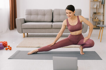 Fototapeta premium Sporty young woman practicing yoga in front of laptop, watching online tutorials.