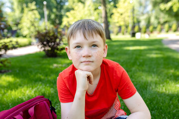portrait of 10 years old schoolboy outdoors. freckled boy sitting on a green lawn in summer. Casual...