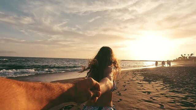 Portrait and close up of young woman holding hand of boyfriend walking on the beach smiling and having fun together. Man following girlfriend to the water of the sea enjoying. 
