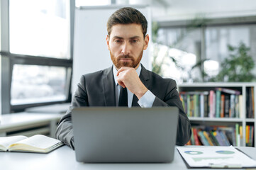 Focused intelligent Caucasian male, marketer, web developer, financial manager, sits at a desktop in front of a laptop, in a modern office in stylish business suit, concentrated working in a laptop