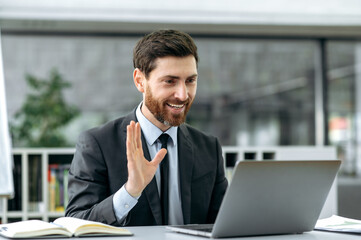 Prospective successful Caucasian male, lawyer, manager, economist, sits at a desk in modern office, wearing stylish business suit, holding video consultation with a client, waving a greeting, smile