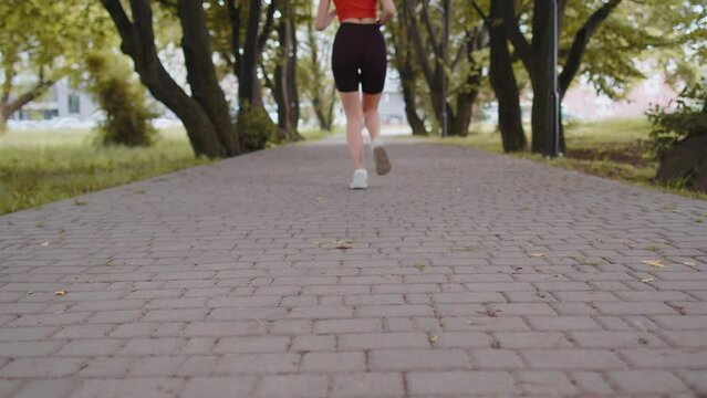 Back view of athletic fit sport jogger girl training marathon run, running through camera on road forward. Workout cardio outdoor in sunny park. Young woman enjoy healthy lifestyle. Active sportswoman