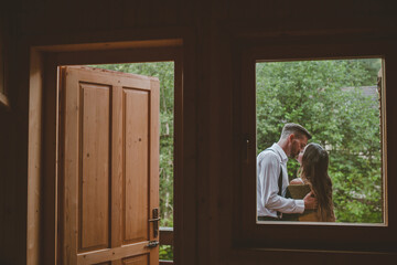 View from inside the cottage. Romantic groom kissing brunette bride on the balcony. Romantic moment. Wedding Day concept